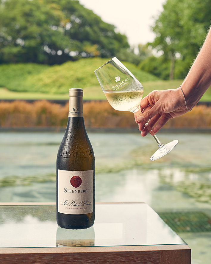 A close-up of a person's hand tilting a glass of Steenberg The Black Swan Sauvignon Blanc, with the elegantly designed bottle on a glass table and a tranquil pond with greenery in the background.