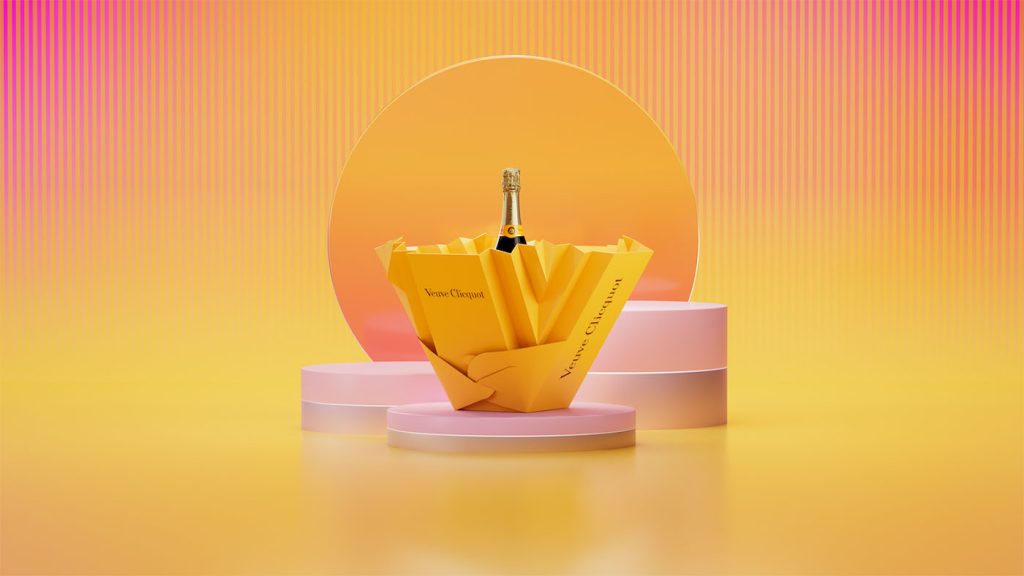 An open Veuve Clicquot Ice Box in yellow, resembling origami art, with a champagne bottle nestled inside, displayed against a pink stand and yellow backdrop.