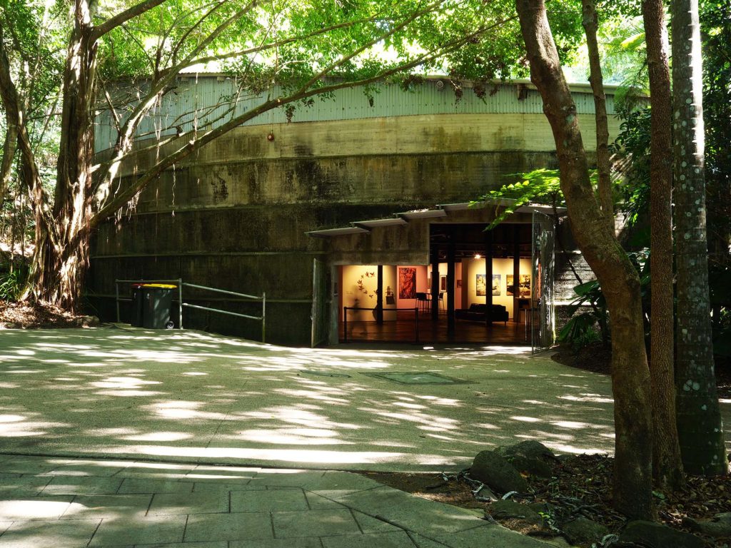 The exterior view of Tanks Arts Centre showing the entrance to the gallery nestled against the one-meter thick concrete walls of a former oil tank, surrounded by the lush greenery of a tropical rainforest.