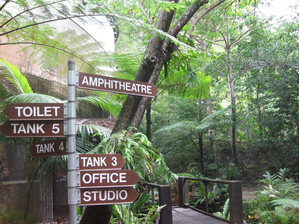 Directional signage at Tanks Arts Centre, pointing to various attractions like the amphitheatre and tanks, set amidst the serene tropical rainforest.