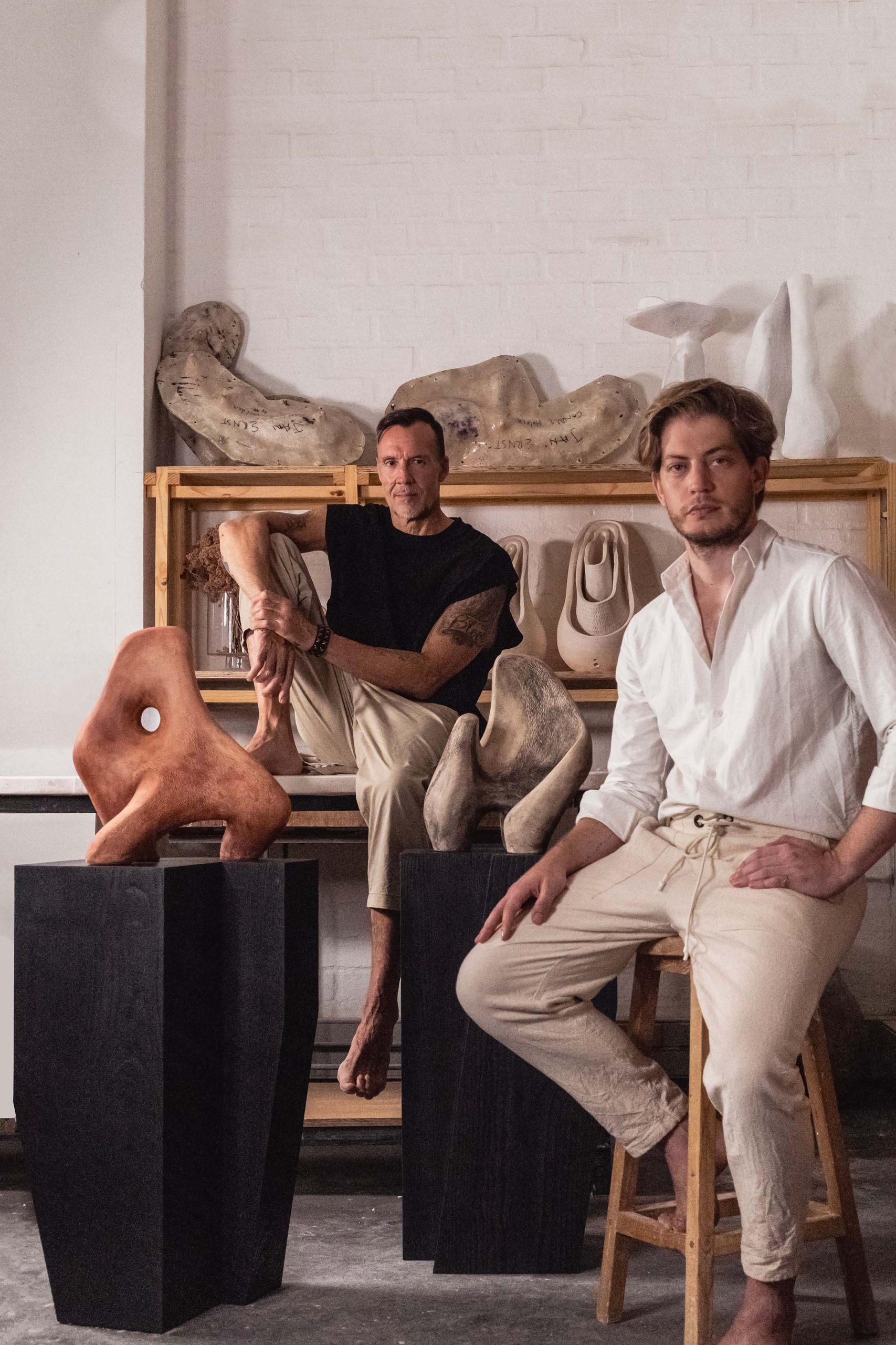 Adam Court and Jan Ernst, captured in a candid moment, are surrounded by their art pieces in a studio setting. Adam leans back on a stool, embodying confidence and creativity, while Jan, seated on a wooden bench, gazes thoughtfully ahead. The background is adorned with a variety of sculptures, illustrating the rich tapestry of talent at OKHA's Cape Town Exhibition, where design and artistry converge.