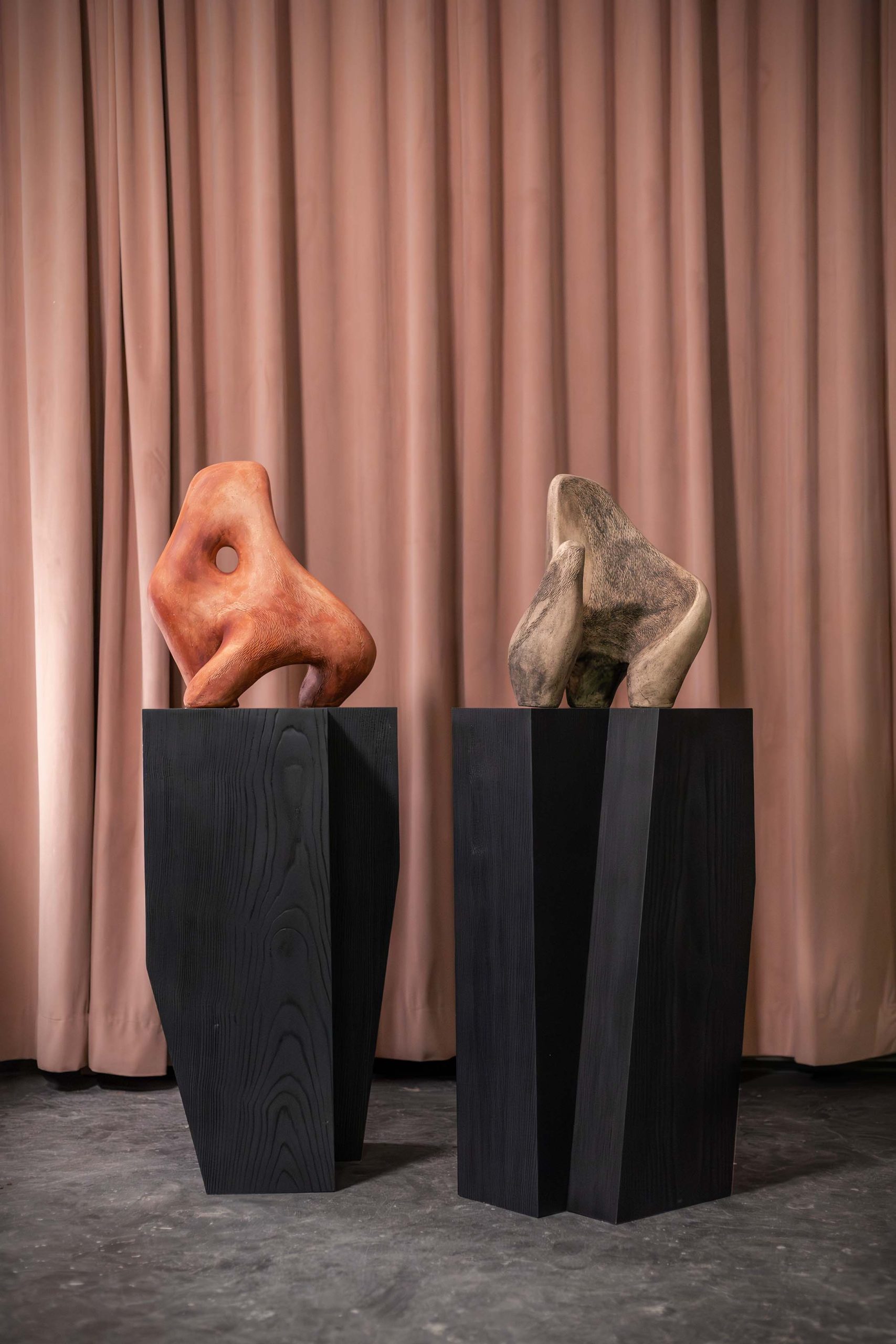 Two abstract sculptures are perched atop angular black plinths, one terracotta-colored with a smooth, curvaceous form and a hollow center, and the other resembling a rough, stone-like texture in neutral hues. These pieces exemplify the fusion of organic and geometric aesthetics, reflecting the innovative design narrative of OKHA's Cape Town Exhibition, set against a backdrop of subtle pink curtains.