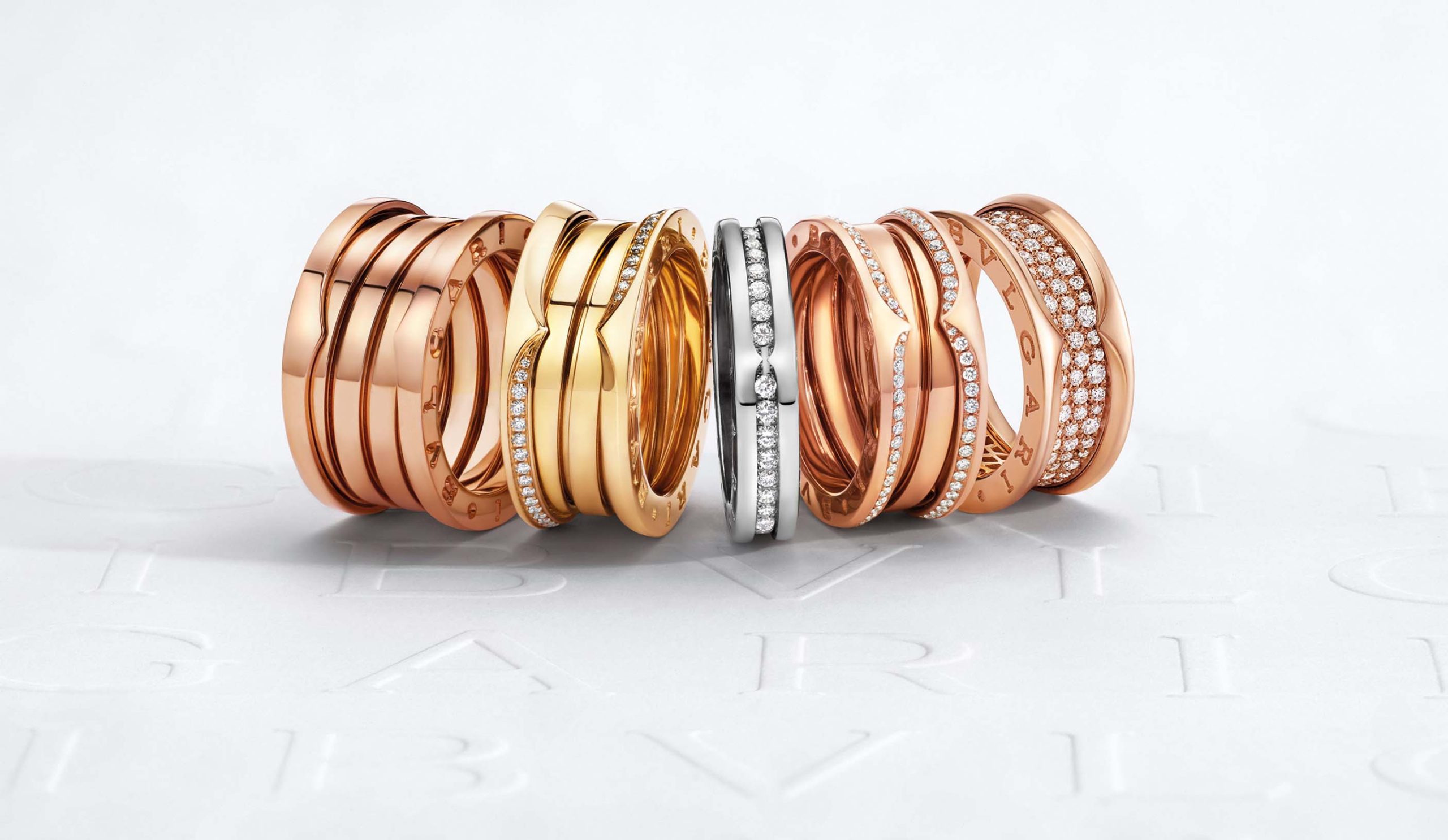 Bulgari Studio: A selection of Bulgari's B.zero1 rings in various designs, featuring a combination of rose gold, yellow gold, and silver bands, some accented with pavé diamonds. The rings are artfully arranged, overlapping each other against a white background with the faint embossed Bulgari logo, showcasing the luxury and craftsmanship of the pieces.