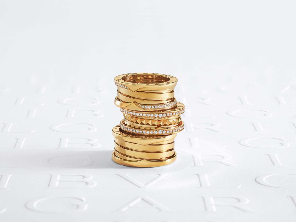 An exquisite stack of Bulgari's B.zero1 yellow gold rings displayed on a white backdrop with a subtle embossed logo, showcasing the intricate design and opulent finish characteristic of Bulgari's luxurious craftsmanship.