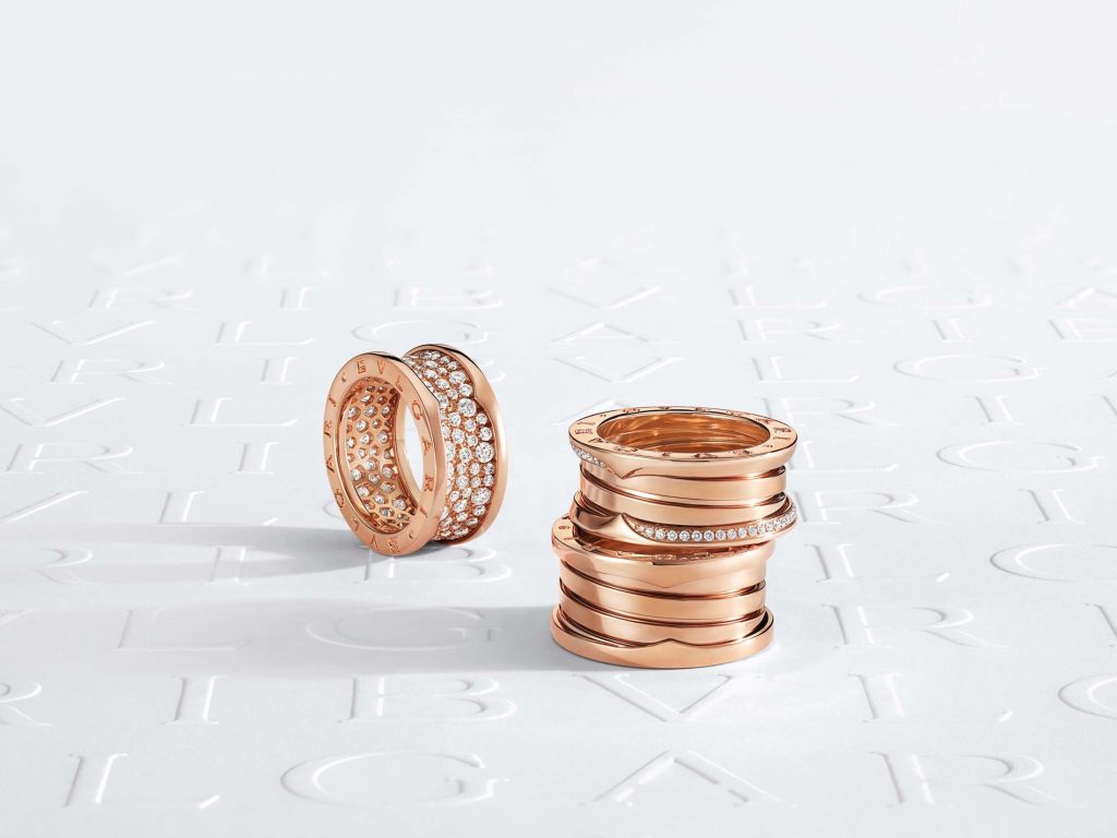 A duo of Bulgari's rose gold B.zero1 rings, one lavishly detailed with diamonds, alongside a neat stack of plain and diamond-studded bands, set against an elegant embossed logo background, illustrating the refined elegance of Bulgari's creations.