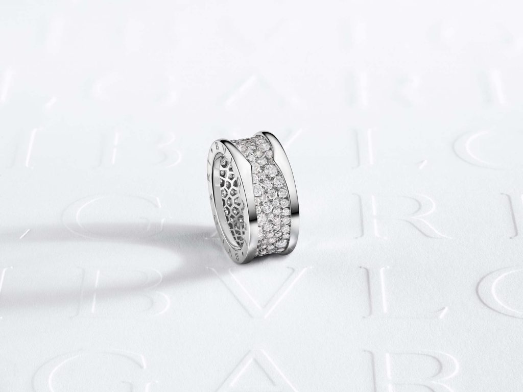 A solitary Bulgari B.zero1 ring in silver, its band partially encrusted with a lavish spread of diamonds, presented on an embossed logo background, capturing the essence of Bulgari's commitment to exceptional quality and artistic design.
