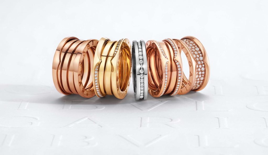 A selection of Bulgari's B.zero1 rings in various designs, featuring a combination of rose gold, yellow gold, and silver bands, some accented with pavé diamonds. The rings are artfully arranged, overlapping each other against a white background with the faint embossed Bulgari logo, showcasing the luxury and craftsmanship of the pieces.A selection of Bulgari's B.zero1 rings in various designs, featuring a combination of rose gold, yellow gold, and silver bands, some accented with pavé diamonds. The rings are artfully arranged, overlapping each other against a white background with the faint embossed Bulgari logo, showcasing the luxury and craftsmanship of the pieces.
