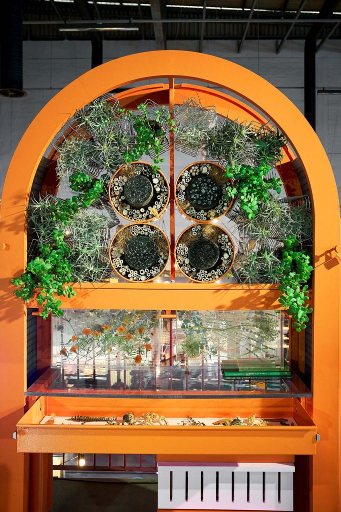 An innovative UPCYCLE display at Maker’s Landing, featuring an orange arched structure with circular cutouts filled with bamboo and greenery, above a piano-key decorated desk.