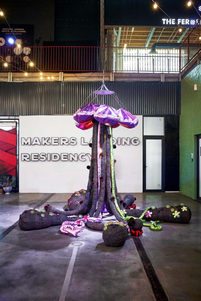 A creative UPCYCLE sculpture at the V&A Waterfront exhibition, resembling a purple tentacled umbrella rising from a base of organic textures and adorned with colorful accents.