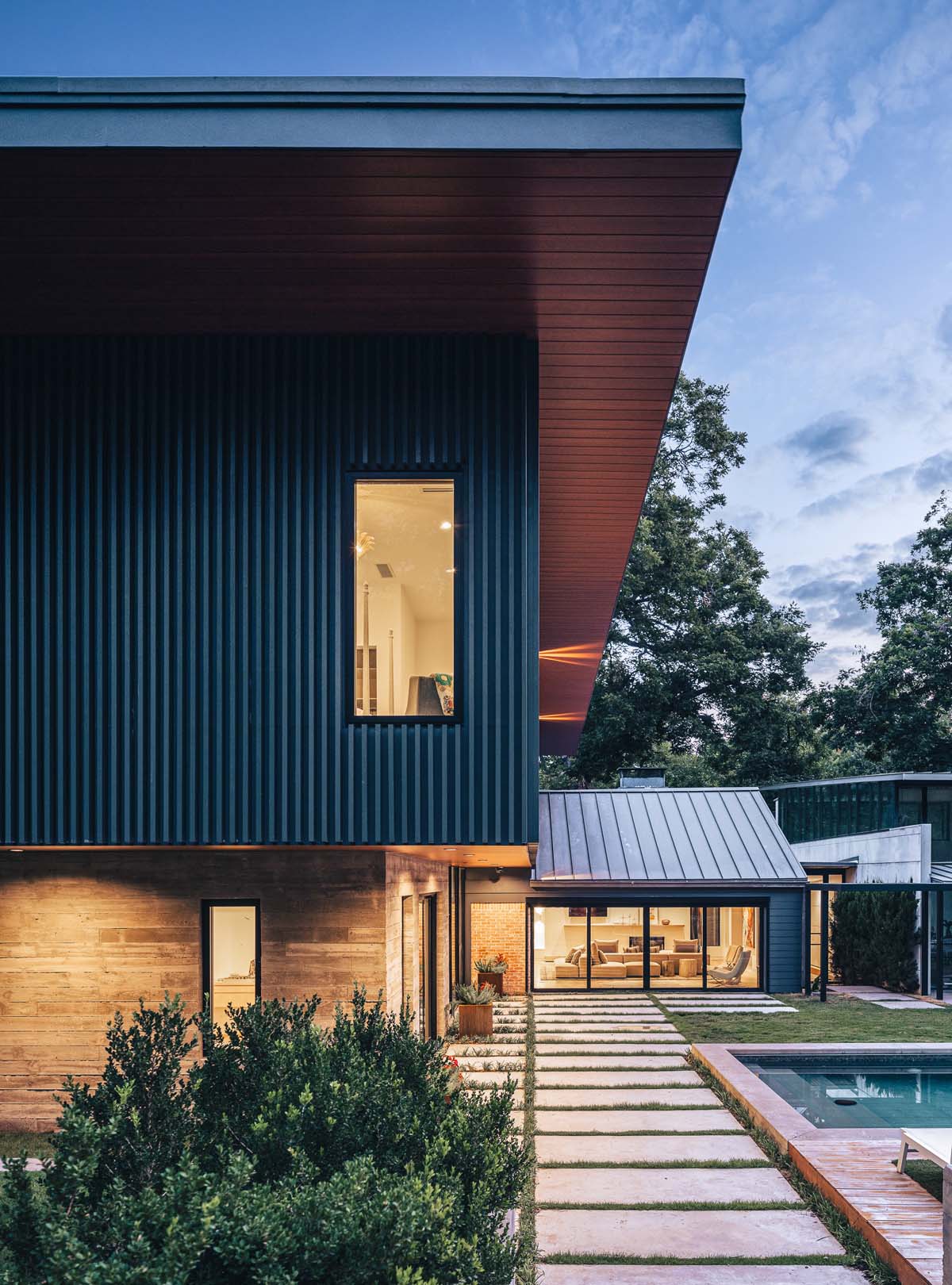 Elegant view of Dissonant House and pool area during twilight, featuring a seamless blend of natural stone, textured battens, and warm interior lighting that enhances the outdoor ambiance.