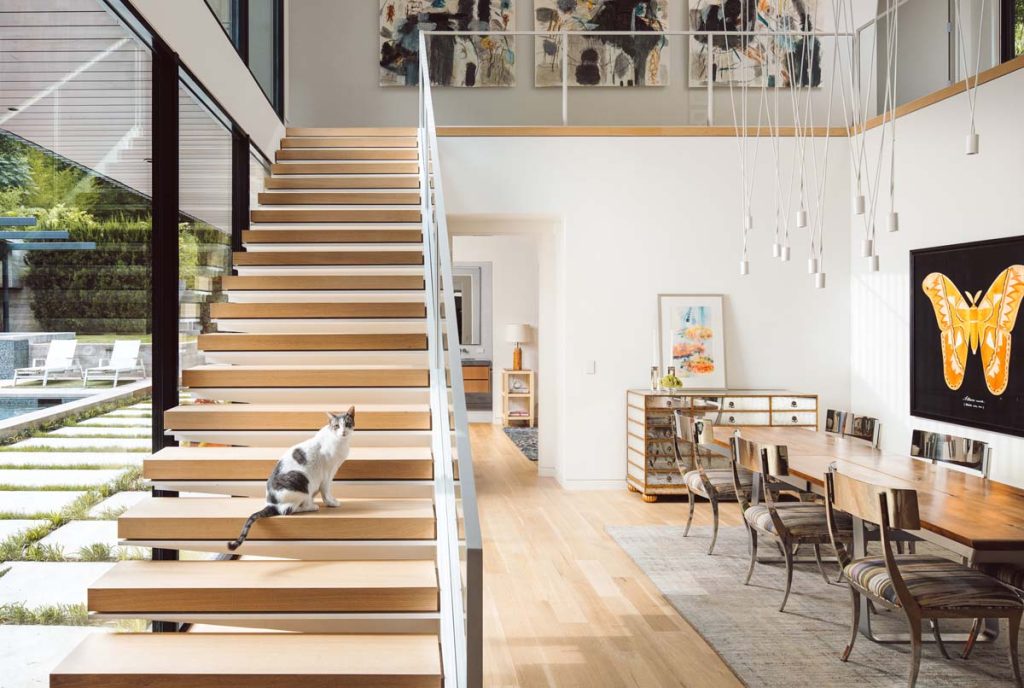 Modern floating staircase in Dissonant House with wooden steps and glass railings, offering a view of the double-height dining area, showcasing the harmonious blend of modern architecture with natural light.