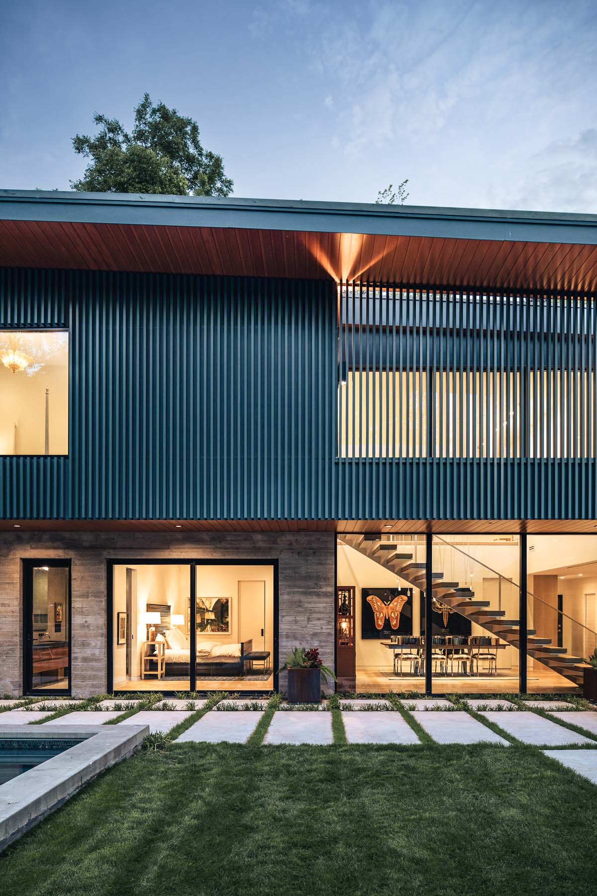 Modern exterior of Dissonant House at dusk, showcasing the rhythmic aluminum battens and large windows that blend into the natural surroundings, creating a seamless indoor-outdoor connection.