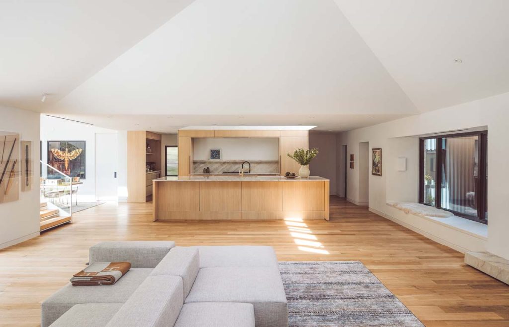 Open-plan kitchen and living area in Dissonant House with light wood flooring, a spacious kitchen island, and natural light streaming through large windows, creating an inviting and modern family space.