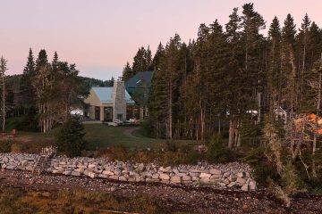 Wide exterior shot of the Belly of the Whale Residence by Acre Architects, showcasing the two distinct volumes against the forest edge and a rocky coastal wall.